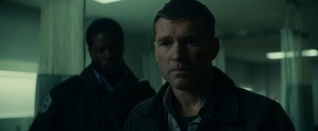Movie still from “Fractured” (2019), directed by Brad Anderson – A man standing next to another man in a dark room; Close Up shot, Low angle