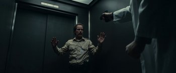 Movie still from “Fractured” (2019), directed by Brad Anderson – A man in a brown shirt is in an elevator with a gun pointed at him; Medium shot, Low angle