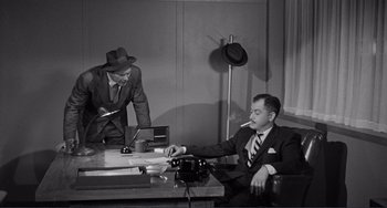 Movie still from “My Gun Is Quick” (1957), directed by Victor Saville – A man sitting at a desk smoking a cigarette while another man stands behind him; Medium shot, Low angle