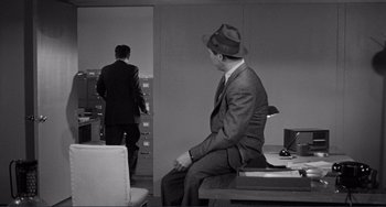 Movie still from “My Gun Is Quick” (1957), directed by Victor Saville – A man in a suit and a hat sitting on a desk; Medium shot, Over the shoulder angle
