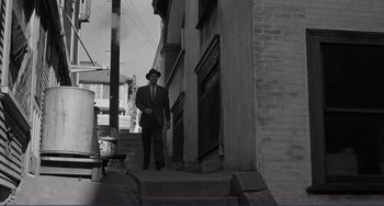 Movie still from “My Gun Is Quick” (1957), directed by Victor Saville – A man in a suit and hat standing on a set of stairs; Wide shot, High angle