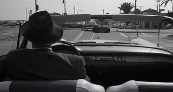 Movie still from “My Gun Is Quick” (1957), directed by Victor Saville – A man in a hat driving a car on a street; Wide shot, Over the shoulder angle