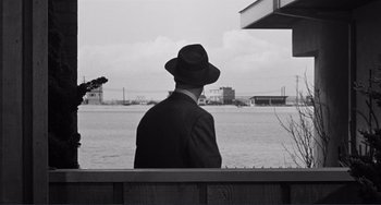 Movie still from “My Gun Is Quick” (1957), directed by Victor Saville – A black and white photo of a man sitting on a bench looking out at a body of water; Medium shot, Low angle