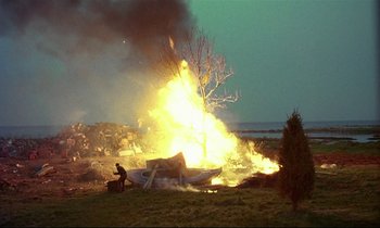 Movie still from “My Life as a Dog” (1985), directed by Lasse Hallström – A man standing in front of a burning boat in a field; Extreme Wide shot, High angle