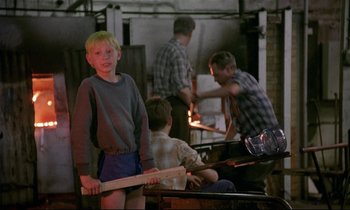 Movie still from “My Life as a Dog” (1985), directed by Lasse Hallström – A young boy holding a baseball bat in front of a group of people; Medium shot, Over the shoulder angle