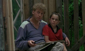 Movie still from “My Life as a Dog” (1985), directed by Lasse Hallström – A man sitting next to a young boy holding a book; Medium shot, High angle