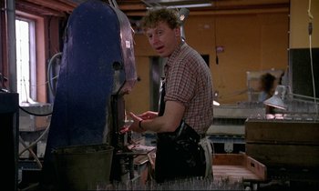 Movie still from “My Life as a Dog” (1985), directed by Lasse Hallström – A man wearing an apron and a checkered shirt is working in a factory; Medium shot, Low angle