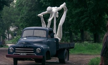 Movie still from “My Life as a Dog” (1985), directed by Lasse Hallström – A truck with a statue of a person in the back of the truck; Wide shot, Low angle