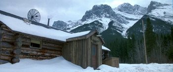 Movie still from “Mystery, Alaska” (1999), directed by Jay Roach – A log cabin in the snow with mountains in the background; Extreme Wide shot, High angle