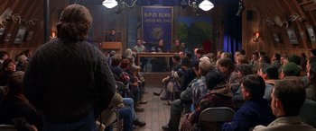 Movie still from “Mystery, Alaska” (1999), directed by Jay Roach – A group of people sitting in front of microphones in a room; Wide shot, High angle