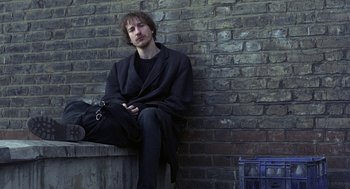 Movie still from “Naked” (1993), directed by Mike Leigh – A man sitting against a brick wall with his hands in his pockets; Medium shot, Low angle