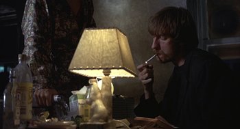 Movie still from “Naked” (1993), directed by Mike Leigh – A man sitting at a table smoking a cigarette next to a lamp; Close Up shot, High angle