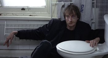 Movie still from “Naked” (1993), directed by Mike Leigh – A man sitting on the floor of a bathroom next to a toilet; Medium shot, High angle