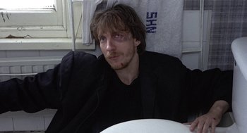 Movie still from “Naked” (1993), directed by Mike Leigh – A man sitting at a table with a towel on the wall behind him; Close Up shot, Low angle