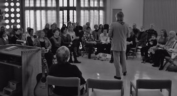Movie still from “Frances Ha” (2012), directed by Noah Baumbach – A man standing in front of a group of people in a room; Wide shot, Over the shoulder angle