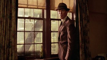 Movie still from “Naked Lunch” (1991), directed by David Cronenberg – A man in a suit and hat standing in front of a window; Medium shot, Low angle