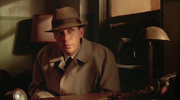 Movie still from “Naked Lunch” (1991), directed by David Cronenberg – A man wearing a hat and a trench coat sitting at a table; Close Up shot, Low angle