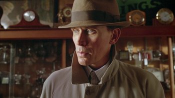 Movie still from “Naked Lunch” (1991), directed by David Cronenberg – A person wearing a hat and a coat; Close Up shot, Over the shoulder angle