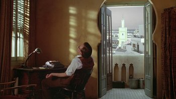 Movie still from “Naked Lunch” (1991), directed by David Cronenberg – A man sitting in a chair in front of an open door; Wide shot, Low angle