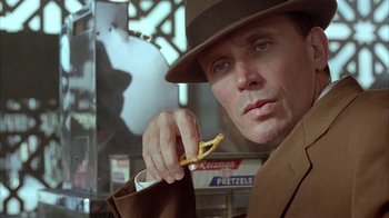 Movie still from “Naked Lunch” (1991), directed by David Cronenberg – A man in a brown suit and hat holding a snack; Close Up shot, Over the shoulder angle
