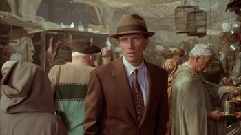 Movie still from “Naked Lunch” (1991), directed by David Cronenberg – A man in a suit and hat standing in front of a group of people; Close Up shot, Over the shoulder angle