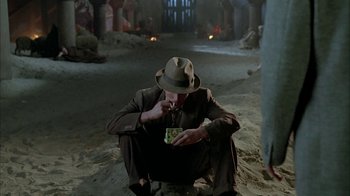 Movie still from “Naked Lunch” (1991), directed by David Cronenberg – A man sitting on the ground with a cigarette in his mouth; Wide shot, High angle