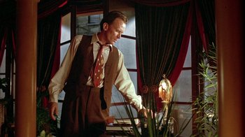 Movie still from “Naked Lunch” (1991), directed by David Cronenberg – A man standing next to a potted plant in front of a window; Medium shot, Low angle