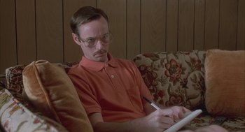 Movie still from “Napoleon Dynamite” (2004), directed by Jared Hess – A man sitting on top of a couch writing on a piece of paper; Medium shot, Low angle