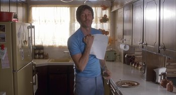 Movie still from “Napoleon Dynamite” (2004), directed by Jared Hess – A man standing in a kitchen holding a piece of paper; Medium shot, Low angle