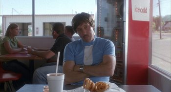 Movie still from “Napoleon Dynamite” (2004), directed by Jared Hess – A man sitting at a table with a drink in front of him; Medium shot, Low angle