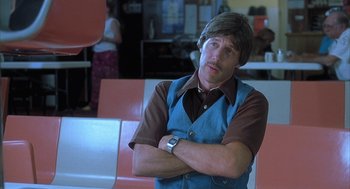Movie still from “Napoleon Dynamite” (2004), directed by Jared Hess – A man with his arms crossed standing in front of a counter; Close Up shot, Low angle