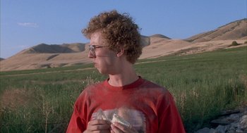 Movie still from “Napoleon Dynamite” (2004), directed by Jared Hess – A person standing in the grass holding a sandwich; Medium shot, Over the shoulder angle