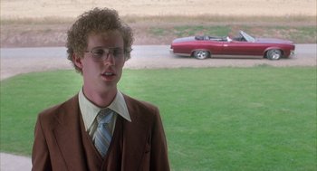 Movie still from “Napoleon Dynamite” (2004), directed by Jared Hess – A man in a suit and tie standing in front of a car; Medium shot, Over the shoulder angle