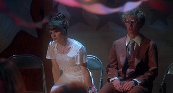 Movie still from “Napoleon Dynamite” (2004), directed by Jared Hess – A woman in a white dress sitting next to a man in a brown suit; Medium shot, Low angle