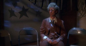 Movie still from “Napoleon Dynamite” (2004), directed by Jared Hess – A man in a suit and tie sitting in front of a chair; Wide shot, Low angle