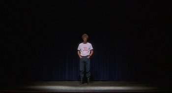 Movie still from “Napoleon Dynamite” (2004), directed by Jared Hess – A man standing on a stage in front of a black curtain; Wide shot, Low angle