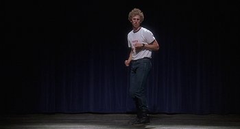 Movie still from “Napoleon Dynamite” (2004), directed by Jared Hess – A man standing on a stage holding a baseball bat; Wide shot, Low angle