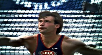 Movie still from “Naqoyqatsi” (2002), directed by Godfrey Reggio – A man in a blue and red tank top is standing in front of a net; Close Up shot, Low angle