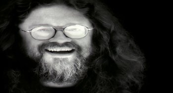 Movie still from “Naqoyqatsi” (2002), directed by Godfrey Reggio – A man with long hair and glasses is smiling for the camera; Extreme Close Up shot, Overhead angle