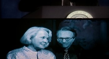 Movie still from “Naqoyqatsi” (2002), directed by Godfrey Reggio – An older man standing next to an older woman in front of an american flag; Close Up shot, Low angle