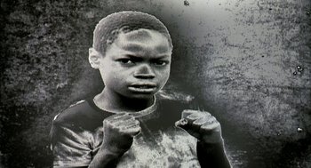 Movie still from “Naqoyqatsi” (2002), directed by Godfrey Reggio – A black and white photo of a young boy with a cigarette in his mouth; Extreme Close Up shot, High angle