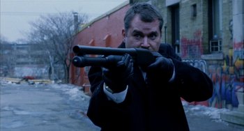Movie still from “Narc” (2002), directed by Joe Carnahan – A man in a black jacket and black gloves is holding a gun; Medium shot, Low angle