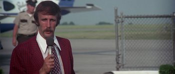 Movie still from “Nashville” (1975), directed by Robert Altman – A man in a red jacket is holding a microphone in front of an airplane on a runway; Medium shot, Low angle