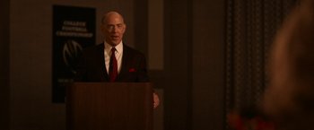 Movie still from “National Champions” (2021), directed by Ric Roman Waugh – A man standing at a podium in front of an audience; Medium shot, Over the shoulder angle