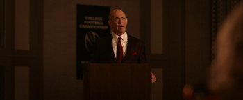 Movie still from “National Champions” (2021), directed by Ric Roman Waugh – A man in a suit and tie standing in front of a podium; Medium shot, Low angle