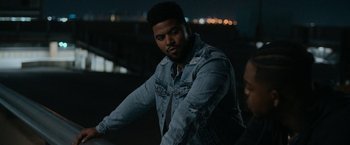 Movie still from “National Champions” (2021), directed by Ric Roman Waugh – A man in a denim jacket standing in the dark; Medium shot, Over the shoulder angle