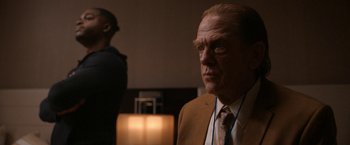 Movie still from “National Champions” (2021), directed by Ric Roman Waugh – A man in a suit and tie standing in front of a lamp; Medium shot, Over the shoulder angle