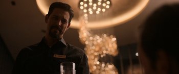 Movie still from “National Champions” (2021), directed by Ric Roman Waugh – A man in a black dress shirt and tie is sitting at a table with a glass; Medium shot, Over the shoulder angle