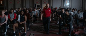 Movie still from “National Champions” (2021), directed by Ric Roman Waugh – A group of people sitting in a room with a man in a red shirt; Wide shot, Over the shoulder angle