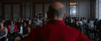 Movie still from “National Champions” (2021), directed by Ric Roman Waugh – A crowd of people sitting in a room with a man wearing a red shirt; Wide shot, Over the shoulder angle